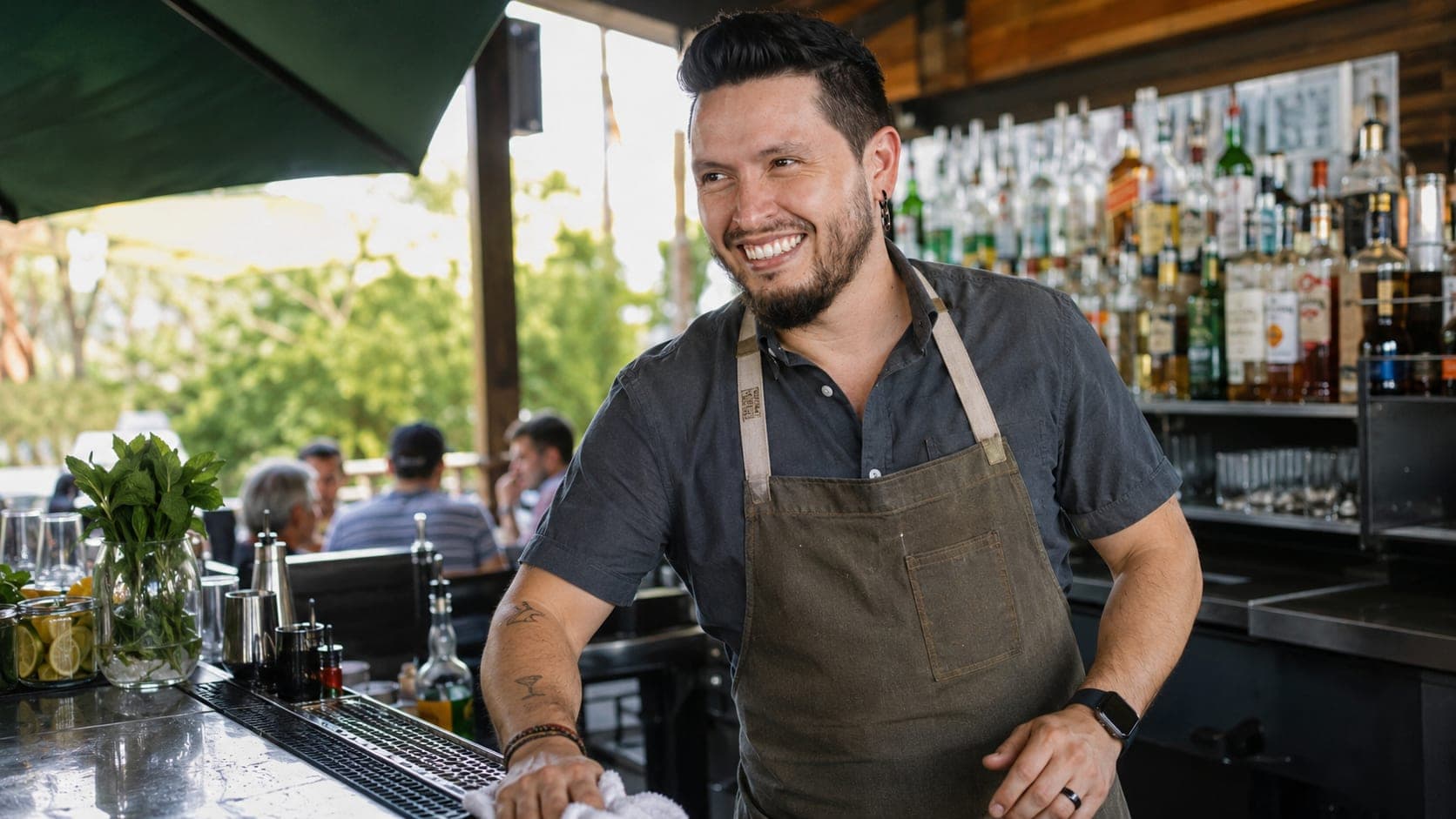 A bartender wipes down an outdoor event bar in daylight, with a few guests seated nearby.